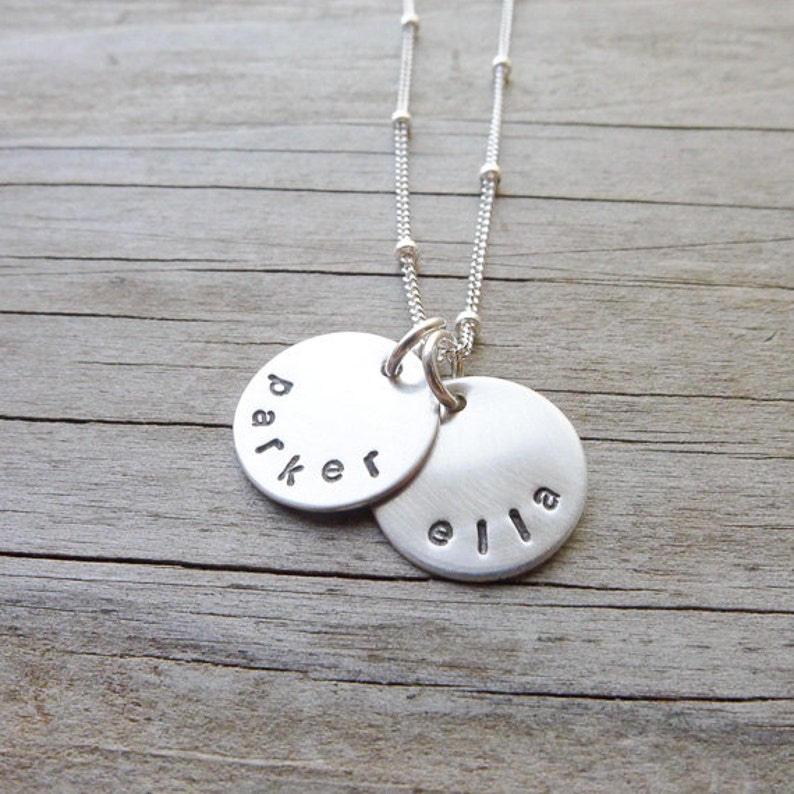May include: Silver necklace with two round pendants. The larger pendant has the name "Parker" engraved on it. The smaller pendant has the name "Ella" engraved on it.