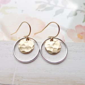 May include: Gold and silver hoop earrings with a hammered gold disc pendant. The earrings have a simple, modern design.