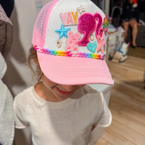 Puede incluir: Gorra de camionero rosa y blanca con la palabra "YAY" y varios parches decorativos. Los parches incluyen una estrella, un osito de peluche y una silueta. La visera es rosa y la malla es blanca y rosa.