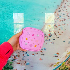 May include: A small pink pouch with a zipper closure. The pouch is decorated with colorful glitter, stars, hearts, and gummy bear shapes.