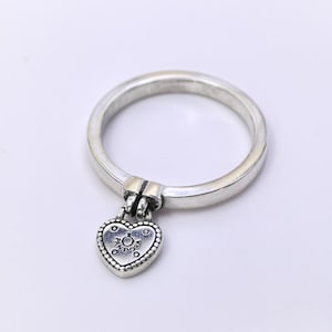 Pode incluir: Um anel de prata com um pingente em forma de coração. O pingente é um design de cadeado com a palavra "Pandora" inscrita. O anel está em um fundo branco.