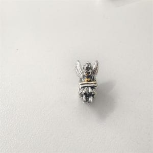 Puede incluir: Un pequeño charm de ángel plateado con alas detalladas y una banda dorada en la cintura. El ángel está sentado, con las manos juntas. El charm mide aproximadamente 1,5 cm de alto y es para una pulsera o collar.