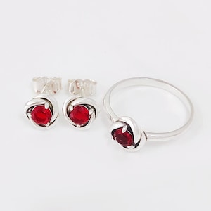 Op de afbeelding: Een zilveren sieradenset met een ring en bijpassende oorknopjes. Elk stuk heeft een knoopontwerp met een heldere rode edelsteen in het midden. De oorbellen hebben een vlindersluiting. De set wordt op een witte achtergrond getoond.