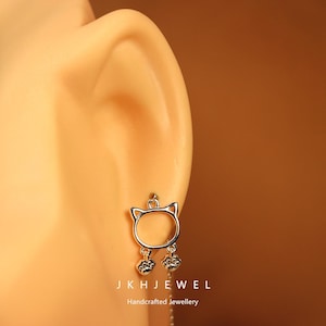 Könnte beinhalten: Silberne Ohrringe in Katzenform mit baumelnden Pfotenabdrücken. Die Ohrringe werden an einem Ohr getragen, mit dem Markennamen "JKH JEWEL" und dem Text "Handcrafted Jewellery" unten.