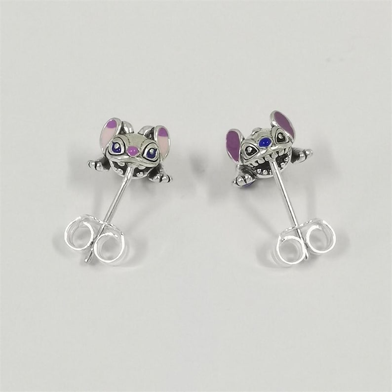May include: A pair of silver stud earrings with a cartoon character design. The earrings show a small, stylized creature with purple ears, blue eyes, and a smiling expression. The earrings have a post and butterfly backing.