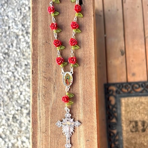 Op de afbeelding: Een rozenkrans met rode rozenkralen en groene bladeren, geregen op een zilveren ketting. Het heeft een zilveren kruisbedel en een kleine ovale bedel met een religieuze afbeelding. De rozenkrans ligt op een houten oppervlak.