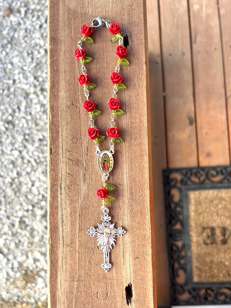 Op de afbeelding: Een rozenkrans met rode rozenkralen en groene bladeren, geregen op een zilveren ketting. Het heeft een zilveren kruisbedel en een kleine ovale bedel met een religieuze afbeelding. De rozenkrans ligt op een houten oppervlak.