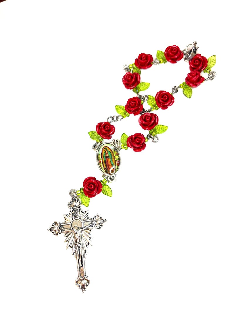 Op de afbeelding: Een rozenkrans met rode rozenkralen en groene bladeren. De rozenkrans heeft een zilveren kruisbeeld en een centrale hanger met een religieuze afbeelding. De kralen zijn verbonden door een zilveren ketting.