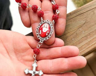 May include: A red beaded rosary with a silver crucifix and a central oval pendant featuring an image of Jesus. The beads are a vibrant red, and the metal components are silver-toned.