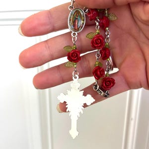 Op de afbeelding: Een rozenkrans met rode rozenkralen, groene bladeren en een zilverkleurige kruisbedel. De rozenkrans bevat een ovale hanger met een religieuze afbeelding.