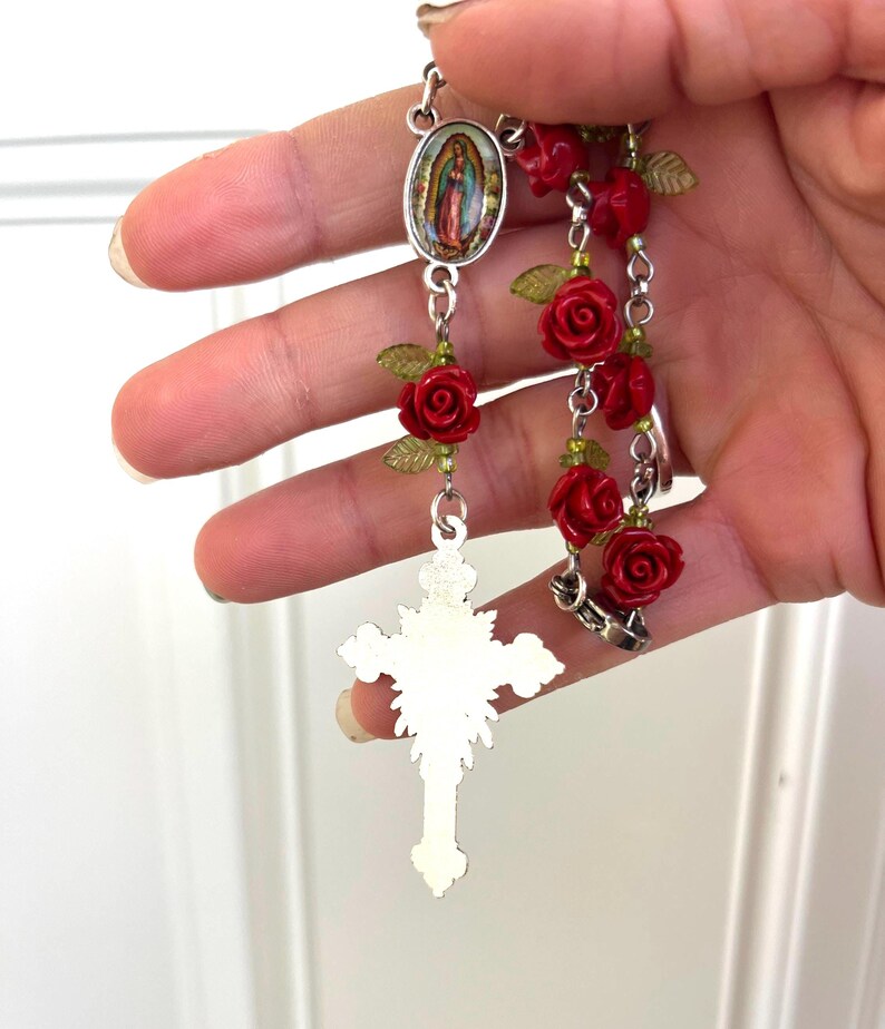 Op de afbeelding: Een rozenkrans met rode rozenkralen, groene bladeren en een zilverkleurige kruisbedel. De rozenkrans bevat een ovale hanger met een religieuze afbeelding.