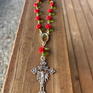 Op de afbeelding: Een rozenkrans met rode rozenkralen en groene bladeren. De rozenkrans heeft een zilveren kruisbeeld met een gedetailleerde afbeelding van Jezus Christus en een centraal medaillon met een religieus beeld.