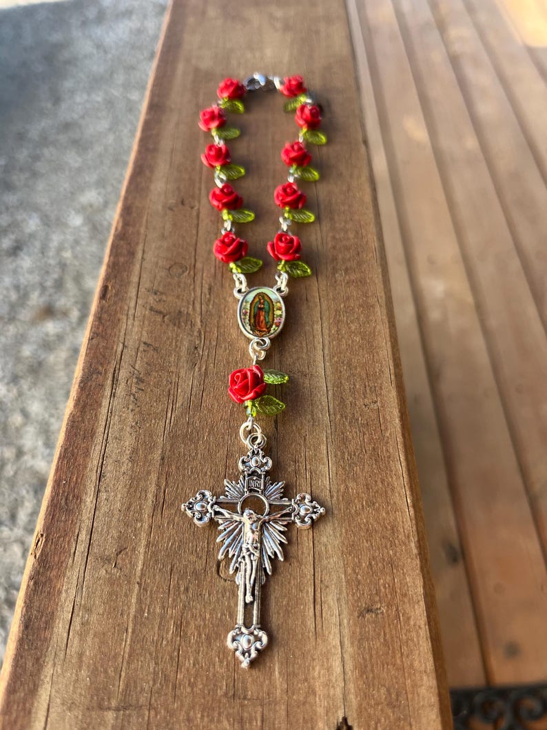 Op de afbeelding: Een rozenkrans met rode rozenkralen en groene bladeren. De rozenkrans heeft een zilveren kruisbeeld met een gedetailleerde afbeelding van Jezus Christus en een centraal medaillon met een religieus beeld.