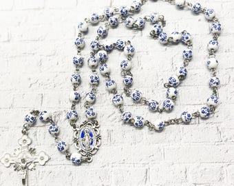 Peut inclure: Un chapelet blanc et bleu avec des perles à motifs floraux. Le chapelet comporte un crucifix argenté et une médaille centrale avec une image bleue. Les perles sont enfilées sur une chaîne argentée.