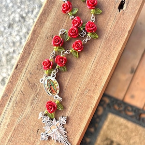 Op de afbeelding: Een rozenkrans met rode rozenkralen en groene bladeren. De rozenkrans heeft een zilveren kruis en een hanger met een religieuze afbeelding. Gepresenteerd op een houten oppervlak.