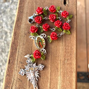 Op de afbeelding: Een rozenkrans met rode rozenkralen en groene bladeren. De rozenkrans heeft een zilveren kruisbeeld en een hanger met een afbeelding van de Maagd Maria. De rozenkrans wordt gepresenteerd op een houten oppervlak.