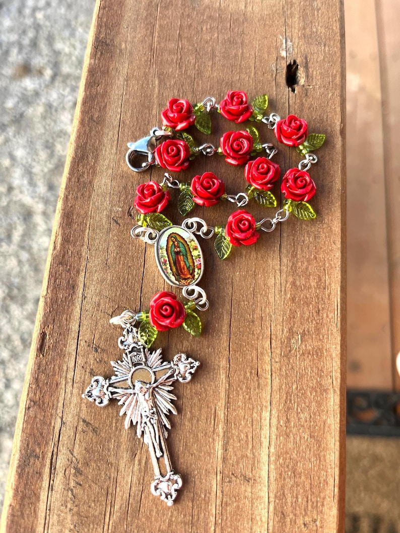 Op de afbeelding: Een rozenkrans met rode rozenkralen en groene bladeren. De rozenkrans heeft een zilveren kruisbeeld en een hanger met een afbeelding van de Maagd Maria. De rozenkrans wordt gepresenteerd op een houten oppervlak.