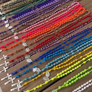 May include: A collection of colorful rosaries displayed on a wooden surface. The rosaries feature knotted cords in various colors, including green, blue, pink, and orange. Each rosary has a silver cross and a medal. The arrangement showcases a variety of religious items.