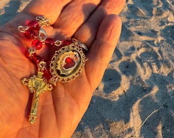 May include: A gold rosary with red beads and a gold crucifix. The rosary features an oval pendant with a religious image. The rosary is held in a hand against a sandy background.