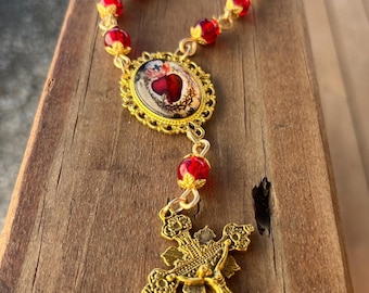 May include: A rosary featuring translucent red glass beads with gold-toned metal caps, a gold-toned oval centrepiece depicting the Sacred Heart of Jesus, and a gold-toned crucifix with an intricate filigree design and a central figure of Christ.