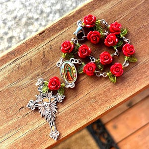 Op de afbeelding: Een rozenkrans met rode rozenkralen en groene bladeren. De rozenkrans heeft een zilveren kruisbeeld met een gedetailleerde afbeelding van Jezus en een kleine ovale hanger met een religieus beeld. De rozenkrans ligt op een houten oppervlak.
