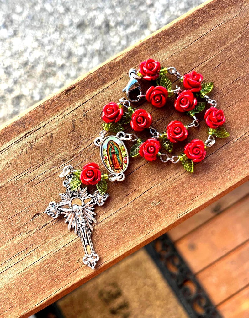 Op de afbeelding: Een rozenkrans met rode rozenkralen en groene bladeren. De rozenkrans heeft een zilveren kruisbeeld met een gedetailleerde afbeelding van Jezus en een kleine ovale hanger met een religieus beeld. De rozenkrans ligt op een houten oppervlak.