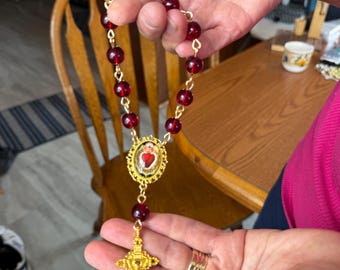 May include: A rosary featuring red beads and a gold-coloured chain and cross. The cross has an intricate design, and the centerpiece displays a heart symbol. The rosary is held in a person's hands.
