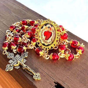 May include: A gold-toned rosary with red beads and a detailed crucifix. The rosary features a central oval pendant with a religious image. The beads are a deep red color, and the metal components have intricate designs.