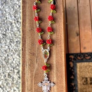 Op de afbeelding: Een rozenkrans met rode rozenkralen en groene bladeren. Het heeft een zilveren kruisbeeld en een hanger met een religieus beeld. De rozenkrans wordt gepresenteerd op een houten oppervlak.