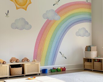 May include: A wall decal set featuring a large watercolor rainbow, a yellow sun, fluffy light blue clouds, and several flying birds. The design is applied to a plain wall above low wooden storage units with woven baskets and wooden toy cars.