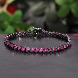 May include: A tennis bracelet featuring a series of round, vibrant pink gemstones set in a dark, black-toned metal frame. The bracelet includes a secure box clasp mechanism, creating a continuous circular design of alternating pink and black.