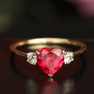 May include: A gold ring featuring a heart-shaped, vibrant red gemstone as the centerpiece, flanked by two smaller, round, clear gemstones. The ring's band is a polished gold color, creating a classic and elegant design.