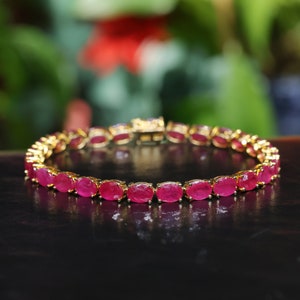 May include: A gold bracelet with a row of oval-shaped ruby gemstones. The bracelet is on a black surface.