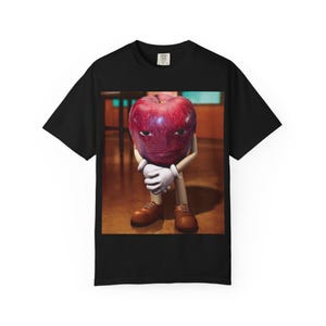 May include: Black t-shirt featuring a cartoon image of a red apple with a face, wearing brown shoes and white gloves. The apple is holding itself. The background is a blurred interior.