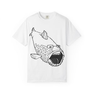 Puede incluir: Camiseta blanca con una ilustración en blanco y negro de un pez con barba, gafas y la boca abierta. El diseño está centrado. La camiseta tiene cuello redondo y mangas cortas.