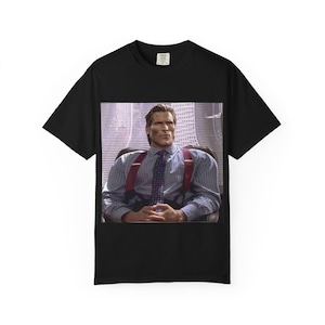 May include: Black t-shirt featuring a printed image of a man in a suit and tie, seated in an office chair. The image on the shirt shows the man wearing a blue striped shirt, red suspenders, and a patterned tie.