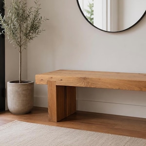 May include: A rectangular wooden bench featuring a thick, natural-toned seat and two sturdy, block-style legs. The bench is crafted from light brown wood with a visible grain pattern, showcasing a simple, functional design with clean, straight lines.