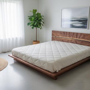 K&ouml;nnte beinhalten: Ein modernes Schlafzimmer mit einem Bett aus Holz und einem Kopfteil aus Naturholz. Das Bett ist mit einer wei&szlig;en, gesteppten Matratze ausgestattet. Ein grauer Sessel, ein runder Jute-Teppich und ein schwarzer Nachttisch mit Lampe sind ebenfalls zu sehen.