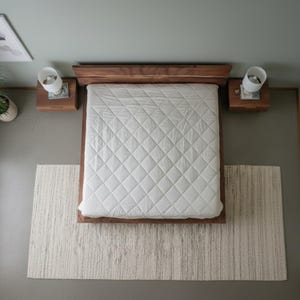 K&ouml;nnte beinhalten: Draufsicht auf ein Schlafzimmer mit einem Holzrahmenbett und einer wei&szlig;en, gesteppten Bettdecke. Zwei Nachttische mit Lampen flankieren das Bett. Ein strukturierter Teppich liegt am Fu&szlig; des Bettes, mit einer Topfpflanze und Kunstwerken.