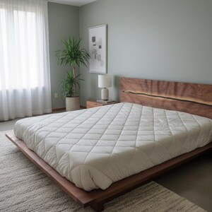 K&ouml;nnte beinhalten: Ein modernes Schlafzimmer mit einem Holzrahmenbett und einem Kopfteil mit nat&uuml;rlicher Kante. Das Bett ist mit einem wei&szlig;en, gesteppten Steppbett ausgestattet. Der Raum hat eine neutrale Farbpalette, mit einem gro&szlig;en Fenster und einer Topfpflanze.