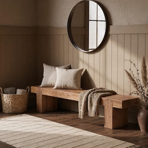 May include: A wooden bench with two beige pillows and a woven throw blanket. A round black framed mirror hangs on the wall above the bench. A woven basket and a vase with dried plants are also in the room. A striped rug is on the floor.