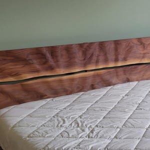 K&ouml;nnte beinhalten: Ein Kopfteil aus massivem Holz mit nat&uuml;rlicher, dunkelbrauner Oberfl&auml;che. Das Kopfteil hat eine einzigartige, organische Form mit einem geteilten Design. Eine wei&szlig;e, gesteppte Matratze ist im Vordergrund zu sehen.