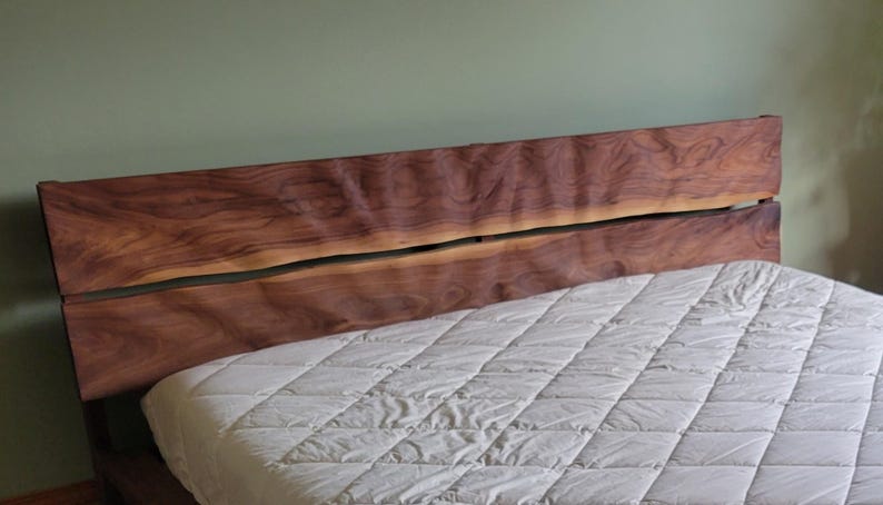 K&ouml;nnte beinhalten: Ein Kopfteil aus massivem Holz mit nat&uuml;rlicher, dunkelbrauner Oberfl&auml;che. Das Kopfteil hat eine einzigartige, organische Form mit einem geteilten Design. Eine wei&szlig;e, gesteppte Matratze ist im Vordergrund zu sehen.