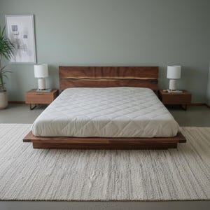 K&ouml;nnte beinhalten: Ein modernes Schlafzimmer mit einem niedrigen Bettrahmen aus Holz und einem Kopfteil aus Naturholz. Das Bett ist mit einer wei&szlig;en, gesteppten Matratze ausgestattet. Zwei passende Nachttische mit Lampen flankieren das Bett. Ein gro&szlig;er Teppich und eine Topfpflanze vervollst&auml;ndigen den Raum.