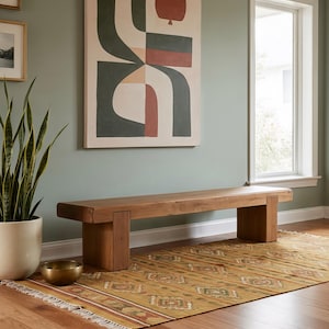 May include: A solid wood bench with a rectangular top and four sturdy legs. The bench is placed on a patterned rug with a fringed edge. An abstract painting hangs on the wall above the bench.
