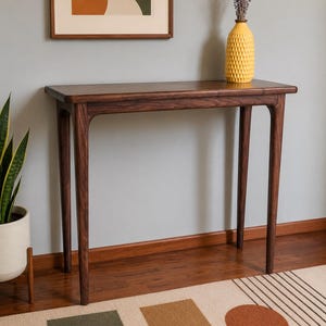 May include: A dark brown wooden console table with a rectangular top and four slender legs. A yellow textured vase with dried lavender sits on the table. A framed abstract art piece hangs on the wall above the table.