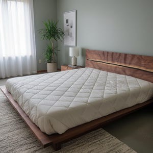K&ouml;nnte beinhalten: Ein modernes Holzbettgestell mit einem Kopfteil aus Naturholz und einer wei&szlig;en, gesteppten Bettdecke. Das Bett steht auf einem hellen Teppich in einem Schlafzimmer mit Fenster und Topfpflanze.