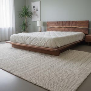 K&ouml;nnte beinhalten: Ein modernes Schlafzimmer mit einem niedrigen Holzrahmenbett und einem Kopfteil aus Naturholz. Das Bett ist mit einer wei&szlig;en, gesteppten Bettdecke bedeckt. Ein cremefarbener Teppich liegt auf dem Boden. Ein Nachttisch mit einer Lampe steht neben dem Bett. Das Bett ist ca. 200 cm lang.