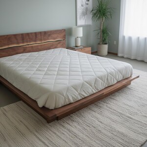 K&ouml;nnte beinhalten: Ein modernes Holzbett mit gro&szlig;em Kopfteil und einer wei&szlig;en, gesteppten Matratze. Das Bett steht auf einem hellen Teppich. Ein Nachttisch mit Lampe und eine Topfpflanze sind zu sehen.