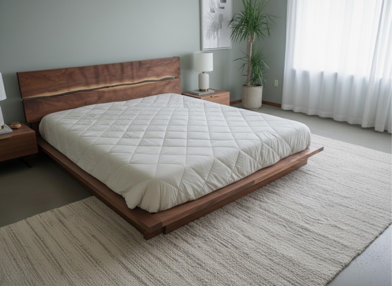 K&ouml;nnte beinhalten: Ein modernes Holzbett mit gro&szlig;em Kopfteil und einer wei&szlig;en, gesteppten Matratze. Das Bett steht auf einem hellen Teppich. Ein Nachttisch mit Lampe und eine Topfpflanze sind zu sehen.